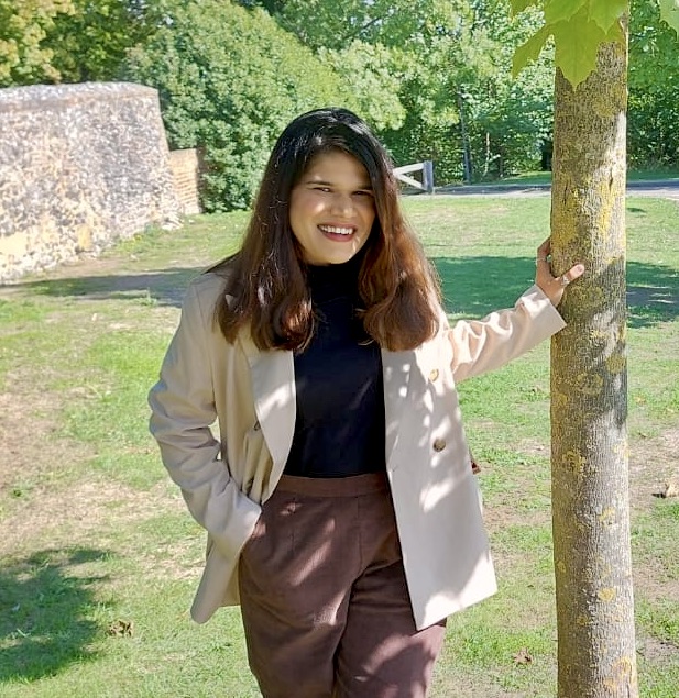 Aparna smiling outdoors, resting a hand on a tree trunk